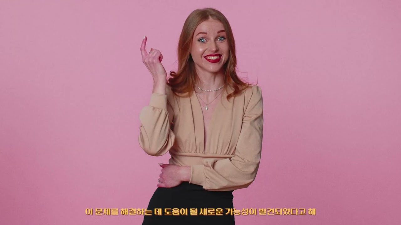 분홍색 배경 앞에서 여성이 미소 지으며 한 손가락을 들어 올리고 화면 아래에 노란 자막이 표시된 장면